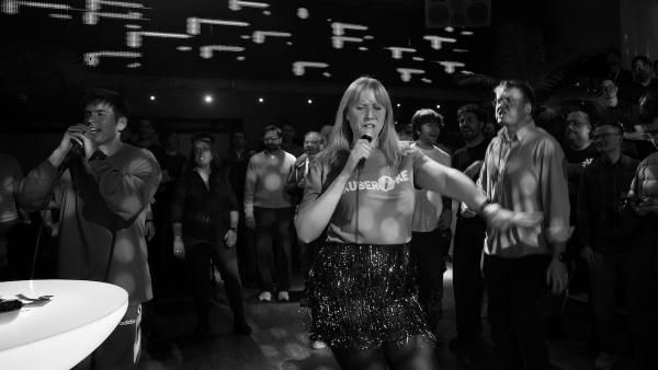 Woman in Kuberoke shirt is standing in the center, singing into a microphone